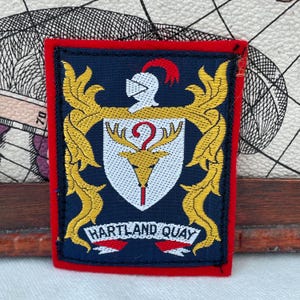 May include: A rectangular patch with a navy blue background and a red border. The patch features a gold emblem with a white shield, a knight's helmet, and the text "HARTLAND QUAY" in white.
