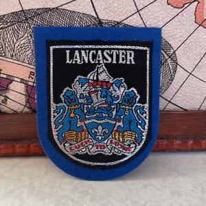 May include: A blue felt patch with a black rectangular center. The center features the word "LANCASTER" in white, above a detailed emblem with a ship, lions, and the text "LUCK TO LOVE". The patch has a silver border.