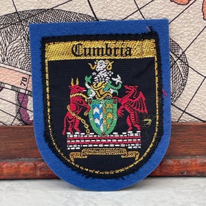 May include: A blue felt patch featuring a black shield with the word Cumbria embroidered in gold at the top. The shield displays a central crest with a red bull and a red dragon on either side, standing on a brick-patterned base with a gold banner below.