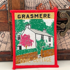 May include: A red, gold and white embroidered patch with the text "Grasmere Dove Cottage". The patch features a white cottage with a pink tree in front of it.