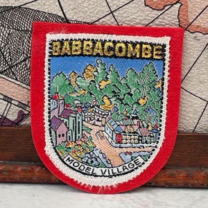 Puede incluir: Un parche bordado en forma de escudo con un borde rojo y costuras blancas. El centro presenta una colorida ilustración paisajística de Babbacombe Model Village con árboles verdes, edificios y un camino. El texto dice BABBACOMBE y MODEL VILLAGE.