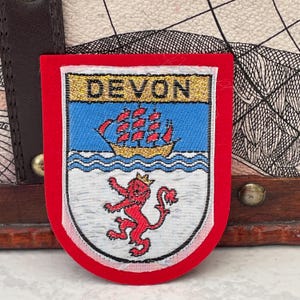 May include: A red-bordered patch featuring the word "DEVON" above a blue and gold emblem. The emblem depicts a ship sailing on wavy lines and a red lion with a crown. The patch is likely a souvenir or emblem.