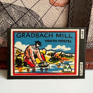 Può includere: Un'etichetta vintage nera e rossa con il testo "Gradbach Mill Youth Hostel" e un'illustrazione di un cartone animato di una persona che cerca l'oro. L'etichetta ha un bordo rosso e un piccolo triangolo verde con la lettera "M" nell'angolo in basso a destra.
