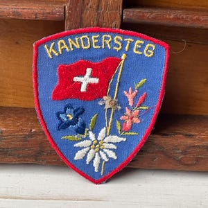 May include: A blue, shield-shaped patch with the word "KANDERSTEG" in yellow. It features a red Swiss flag, flowers, and a white edelweiss. The patch is likely a souvenir or emblem.
