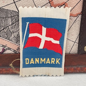 May include: A rectangular fabric patch featuring the flag of Denmark. The flag is red with a white Scandinavian cross on a blue background. The word "DANMARK" is printed in yellow letters below the flag.