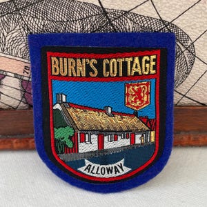 May include: A blue felt patch with the words "BURN'S COTTAGE" in gold lettering. The patch features an illustration of a white cottage with a thatched roof and the word "ALLOWAY" at the bottom.