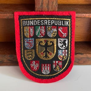 Könnte beinhalten: Ein schildförmiger gestickter Aufnäher mit einem leuchtend roten Filzrand und einem schwarzen Zentrum. Der Text BUNDESREPUBLIK ist oben in Weiß gestickt. Er zeigt ein zentrales schwarzes Adleremblem, umgeben von zehn kleineren bunten Wappenschilden.