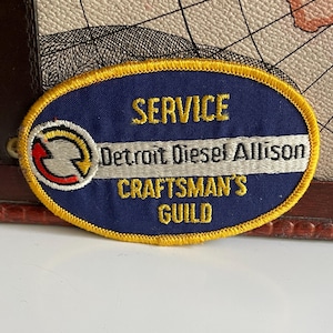 Op de afbeelding: Ovale patch met een marineblauwe achtergrond en gouden rand. De patch bevat de woorden "SERVICE", "Detroit Diesel Allison" en "CRAFTSMAN'S GUILD" in gouden letters. Een witte horizontale streep verdeelt de tekst.