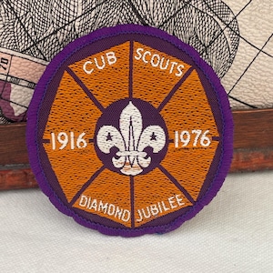 Może przedstawiać: Okrągła naszywka Cub Scouts Diamond Jubilee. Naszywka ma fioletową obwódkę, pomarańczowy środek i biały napis z napisem "CUB SCOUTS", "1916", "1976" i "DIAMOND JUBILEE". Biały fleur-de-lis znajduje się na środku.