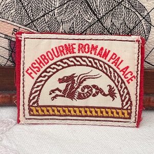 May include: A rectangular patch featuring the text "FISHBOURNE ROMAN PALACE" in red, above a brown dragon illustration. The patch has a cream background, a red border, and a decorative yellow and brown border.
