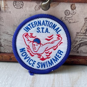 May include: A circular embroidered patch with a blue border. The patch features the words "INTERNATIONAL S.T.A. NOVICE SWIMMER" in blue text and a swimmer in red and white, with a blue swim cap.