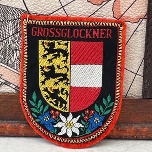 Könnte beinhalten: Ein schildförmiger, bestickter Aufnäher mit leuchtend orangefarbenem Rand. Auf schwarzem Grund steht der rote Schriftzug GROSSGLOCKNER über einem geteilten Wappen: eine gelbe Seite mit schwarzen Löwen und eine rot-weiß-rote Seite. Blaue und weiße Blumen.