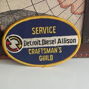 Op de afbeelding: Een ovaalvormige patch met een marineblauwe achtergrond en gouden rand. De patch bevat de woorden "SERVICE", "Detroit Diesel Allison" en "CRAFTSMAN'S GUILD" in gouden letters. Een cirkelvormig logo met een rode en gele pijl staat links.