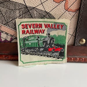 Può includere: Una toppa rettangolare con la scritta "SEVERN VALLEY RAILWAY" in lettere rosse. Sotto, un'illustrazione verde e nera di una locomotiva a vapore. La toppa ha un bordo verde su sfondo color crema.