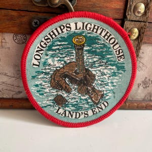 May include: Round patch with a red border featuring a lighthouse scene. The patch has the words "LONGSHIPS LIGHTHOUSE" and "LAND'S END" in white text. The lighthouse is brown and sits on a rocky island, with a blue and green ocean background.