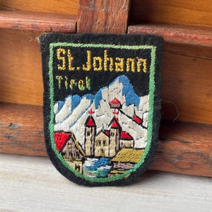 Peut inclure: Écusson brodé avec le texte "St. Johann Tirol" et une scène de montagne et d'église. L'écusson a un fond noir avec une bordure verte. Les montagnes sont blanches et bleues, et l'église a des toits rouges.