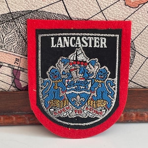 May include: A shield-shaped embroidered patch with a red felt border and a black center. The patch features the word LANCASTER in white text above a detailed crest with two blue lions, a ship, and a blue shield with a white fleur-de-lis.