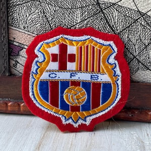 Puede incluir: Un parche bordado en rojo, amarillo y azul con el escudo del FC Barcelona. El diseño del escudo incluye una cruz roja sobre fondo blanco, franjas verticales amarillas y rojas, y las letras C.F.B. en azul sobre una banda horizontal blanca.