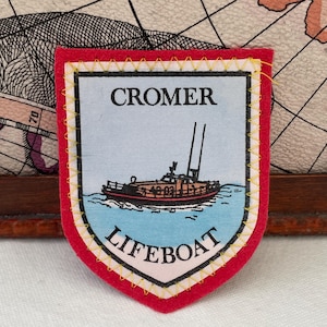 Op de afbeelding: Rood, schildvormig embleem met de tekst "CROMER LIFEBOAT" en een bootillustratie. Het heeft een witte rand met gele stiksels. De achtergrond toont een kaart. Een souvenir.