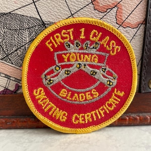 Pode incluir: Um patch redondo vermelho com borda dourada e texto. O patch diz "FIRST 1 CLASS YOUNG BLADES SKATING CERTIFICATE" em letras douradas. O centro apresenta um emblema prateado.