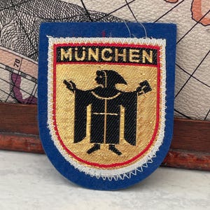 Puede incluir: Parche azul en forma de escudo con la palabra "MUNCHEN" en letras doradas. El parche presenta una figura bordada en oro y negro con los brazos extendidos. El borde es rojo y blanco, con un reverso de fieltro azul.
