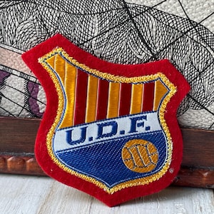 Op de afbeelding: Een schildvormige geborduurde patch met een rode vilten rand. Het bovenste gedeelte heeft verticale gele en rode strepen. Een witte horizontale band in het midden toont de blauwe tekst U.D.F. Een blauw onderste gedeelte bevat een gele voetbal.