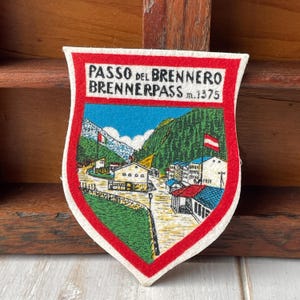 Peut inclure: Un écusson vintage en forme de bouclier avec une bordure rouge et une scène de paysage détaillée. L'écusson porte l'inscription "PASSO DEL BRENNERO BRENNERPASS m.1375" et représente un village de montagne avec des drapeaux, des arbres et des bâtiments.