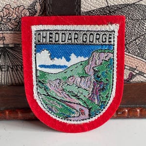 May include: A rectangular patch with a red border and the words "CHEDDAR GORGE" at the top. The patch features a landscape scene with a blue sky, green hills, and a winding road. The patch is likely a souvenir.