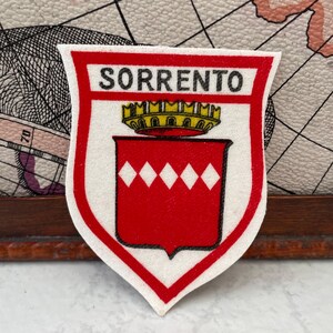 Puede incluir: Un parche de tela en forma de escudo con fondo blanco y borde rojo. El texto SORRENTO está impreso en negro en la parte superior. Debajo del texto hay una corona amarilla sobre un escudo rojo con una fila horizontal de cinco rombos blancos.