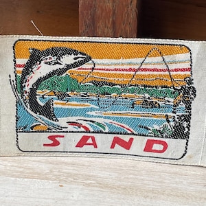 May include: A rectangular fabric patch with a vibrant illustration of a fish leaping from water. The design includes a river scene and the word "SAND" in red. The background is a mix of yellow, green, blue, and red.