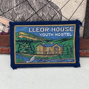 Puede incluir: Parche bordado con el texto "LLEDOR HOUSE YOUTH HOSTEL". El parche presenta un paisaje detallado con un edificio, un río y montañas en tonos verdes, azules y dorados, sobre un borde azul oscuro.