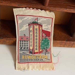 May include: A vintage textile souvenir featuring a detailed image of Rundetårn in Copenhagen, Denmark. The design includes a tall, multi-story tower with a flag, adjacent buildings, and a tree, all framed by a red border. The text "RUNDETÅRN KOBENHAVN" is visible.