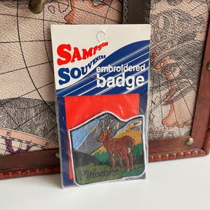 Może przedstawiać: Bordowana odznaka w kolorze czerwonym, białym i niebieskim z tekstem "Sampson Souvenir embroidered badge". Odznaka przedstawia jelenia w górskim krajobrazie z tekstem "Pitlochry" pod jeleniem.
