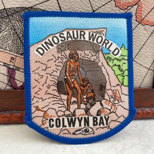 May include: A blue-edged patch with the text "DINOSAUR WORLD" and "COLWYN BAY." The patch shows two figures in front of a cave, with a blue sky and green foliage. The patch has a light brown and blue colour scheme.