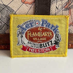 May include: A rectangular yellow embroidered patch featuring the text Cornwall AeroPark and Flambards Village Britain in the Blitz Helston. The design includes illustrations of two figures in period clothing on a white background with red accents.
