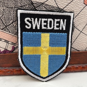 May include: Embroidered patch with the word "SWEDEN" in white on a black background. The patch features the Swedish flag: a blue field with a yellow cross. The patch is edged with white stitching.