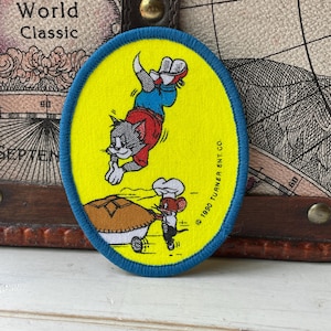 May include: An oval-shaped embroidered patch with a bright yellow background and a blue border. It features a cartoon cat in a red shirt and blue pants jumping toward a brown pie held by a small mouse wearing a white chef hat. Text reads 1990 TURNER ENT. CO.