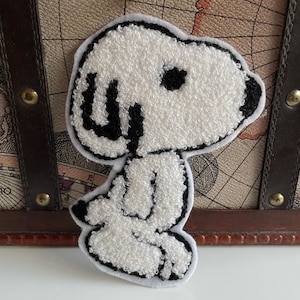 Könnte beinhalten: Ein weiß-schwarzer Snoopy-Aufnäher mit strukturiertem, plüschigem Aussehen. Der Aufnäher hat eine schwarze Umrandung und Details, einschließlich Snoopy's Ohren, Augen und Gesichtszüge. Der Aufnäher ist wahrscheinlich für Kleidung oder Accessoires.