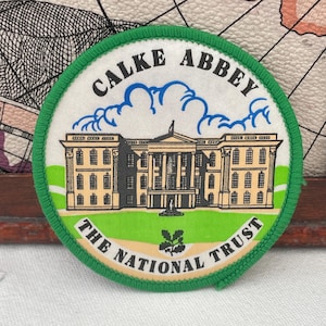 May include: A circular patch with a green border. The patch features the text "CALKE ABBEY" above an illustration of a building. Below the building is the text "THE NATIONAL TRUST". The patch has a white background and a green border.