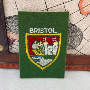 May include: A green patch with the word "BRISTOL" embroidered in white. The patch features a shield design with a red background, a yellow border, and a detailed emblem of a ship, castle, and water.