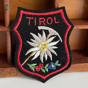 Könnte beinhalten: Ein schwarzer, schildförmiger Aufnäher mit roter Umrandung und dem Wort "TIROL" in Rot gestickt. Der Aufnäher zeigt eine weiße und gelbe Blume, eine Spitzhacke und bunte florale Akzente. Der Aufnäher ist wahrscheinlich ein Souvenir oder ein Emblem.