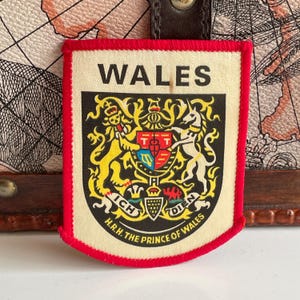 May include: A rectangular patch with a red border and the word "WALES" at the top. The patch features a detailed coat of arms with a shield, a crown, and the text "H.R.H. THE PRINCE OF WALES". The background is a cream color.