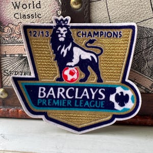 May include: A fabric patch featuring the Barclays Premier League 12/13 Champions logo. The design includes a dark blue lion wearing a crown, standing over a red and white football, set against a gold background with the text Barclays Premier League.