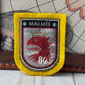 Puede incluir: Un parche bordado rectangular con un borde de fieltro amarillo brillante. El centro presenta un escudo plateado con una cabeza de grifo roja con una corona dorada, las letras BG y el texto MALMÖ en la parte superior flanqueado por dos estrellas.