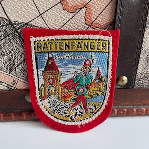 May include: Embroidered patch with the text "Rattenfanger von Hameln" depicting a colourful illustration of a figure playing a flute in front of a castle. The patch has a red border and is set against a map-patterned background.