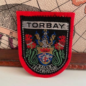 May include: A shield-shaped patch with a red border and black backing. The patch features the word "TORBAY" above a detailed emblem with a crown, two red griffins, and the Latin phrase "SALUS ET FELICITAS".