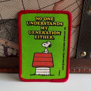 May include: A rectangular patch with a green background and a red border. The patch features Snoopy on top of his doghouse, with the text "NO ONE UNDERSTANDS MY GENERATION EITHER." The patch is a vintage item.