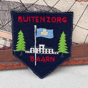 Könnte beinhalten: Marineblauer Aufnäher mit dem roten Text "BUITENZORG BAARN". Der Aufnäher zeigt eine hellblaue Flagge mit gelbem Emblem, zwei grüne Kiefern und ein weißes Gebäude.