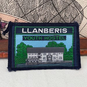 Puede incluir: Un parche rectangular con el texto "LLANBERIS YOUTH HOSTEL". El parche presenta una ilustración de un edificio con árboles y un fondo de cielo azul, enmarcado por un borde azul oscuro. Un recuerdo.