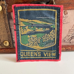 Op de afbeelding: Een rechthoekige patch met een rode rand en een donkerblauw midden. De patch toont een landschap in goud en groen, met de tekst "QUEEN'S VIEW" in gouden letters.